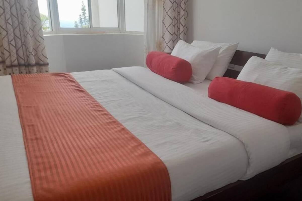 Un lit bien fait, avec une literie blanche et orange, deux oreillers rouges et une fenêtre avec des rideaux.