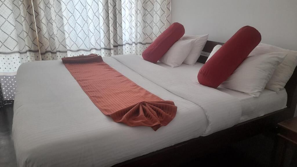 Un lit bien fait, avec des draps blancs et des oreillers rouges, une couverture marron et une fenêtre ornée de rideaux à motifs.