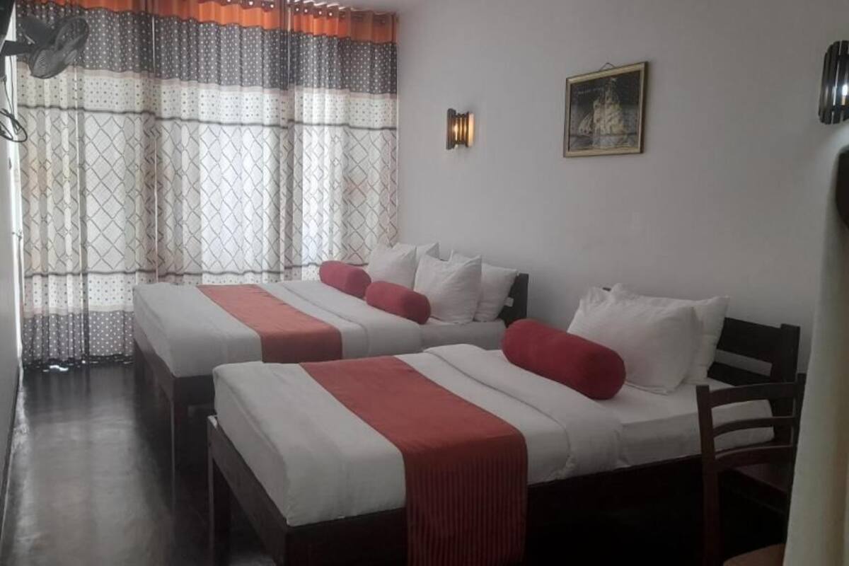 Une chambre d’hôtel avec deux lits, chacun agrémenté de draps blancs et de touches de couleur rouge, une grande fenêtre avec des rideaux à motifs, un luminaire fixé au mur et un tableau encadré sur le mur.
