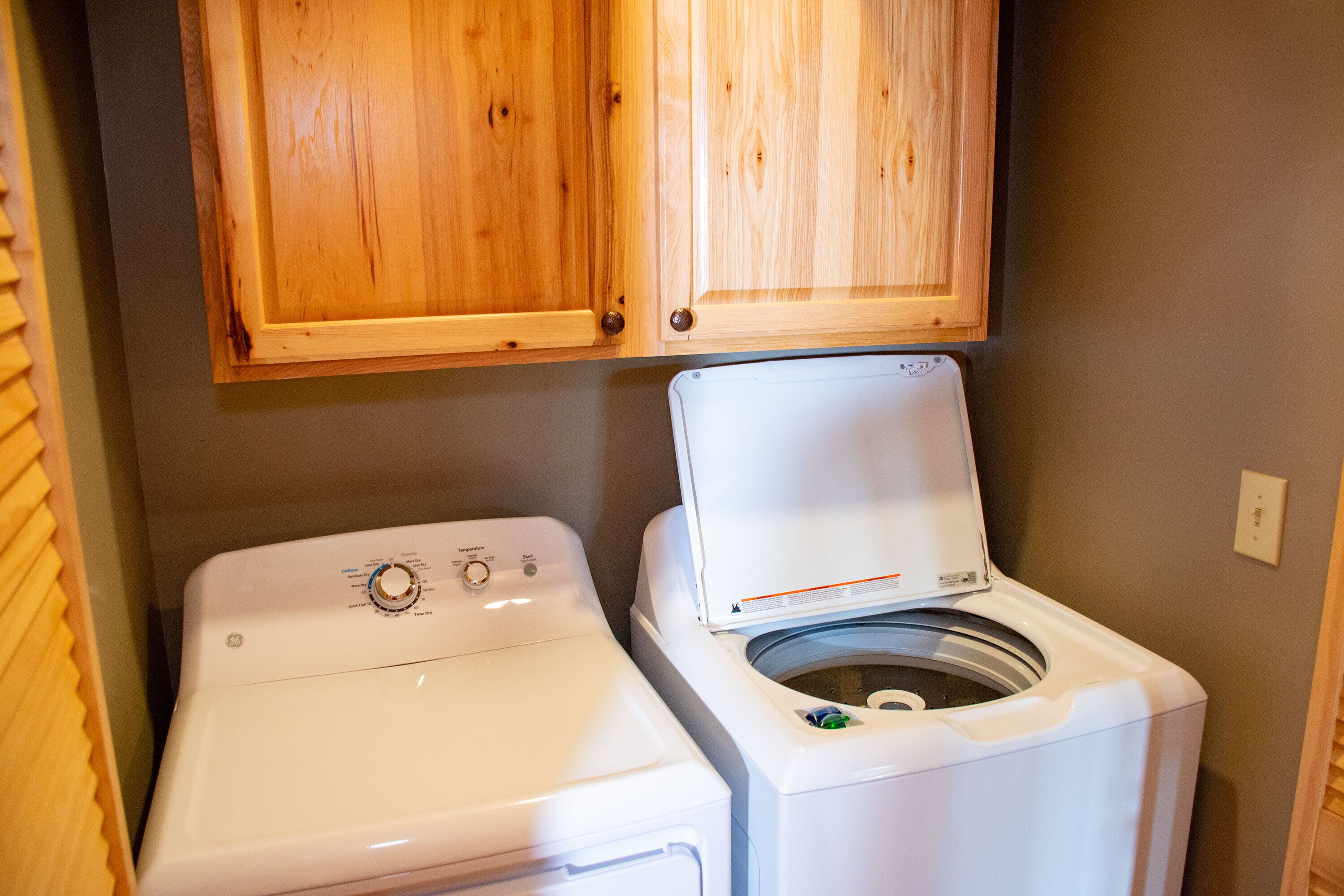 Een wasserette met een voorlader wasmachine en droger, houten kastjes erboven en een grijze wand.