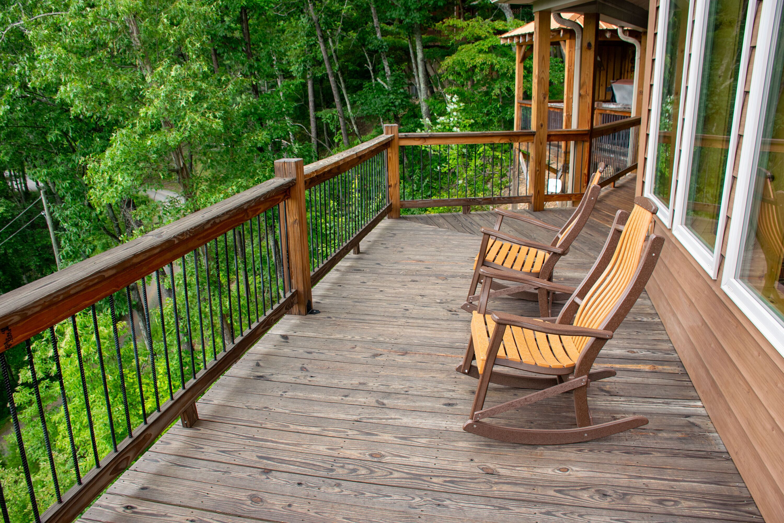A wooden deck with rocking chairs and a glass railing overlooking a forested area.