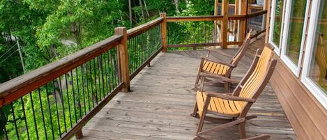 A wooden deck with rocking chairs and a glass railing overlooking a forested area.