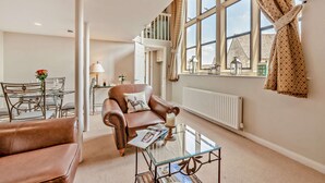 Ein geräumiges Wohnzimmer mit einer Ledersofa, einem Glastisch und einem Essbereich mit Tisch und Stühlen. Große Fenster mit Vorhängen sorgen für natürliches Licht und einen Ausblick nach draußen.