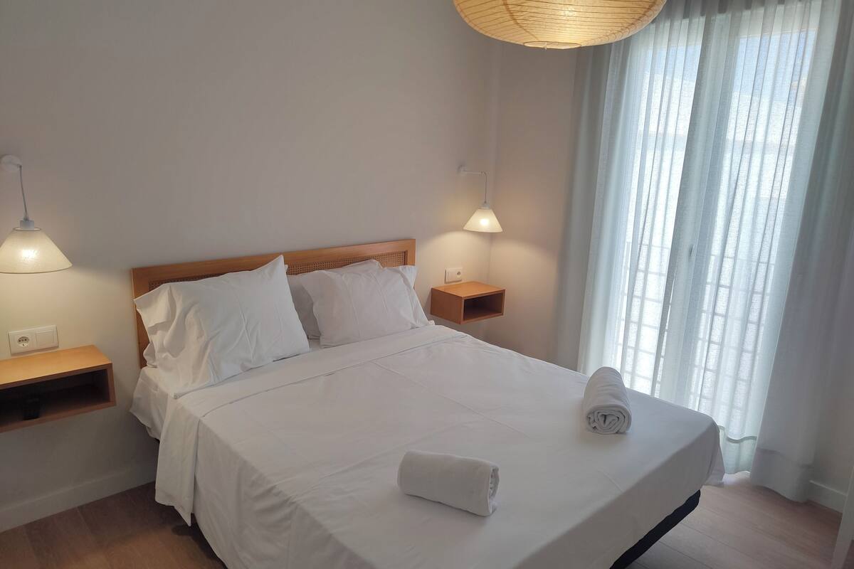 A neatly made bed with white linens, two bedside tables with lamps, and a large window with sheer curtains.