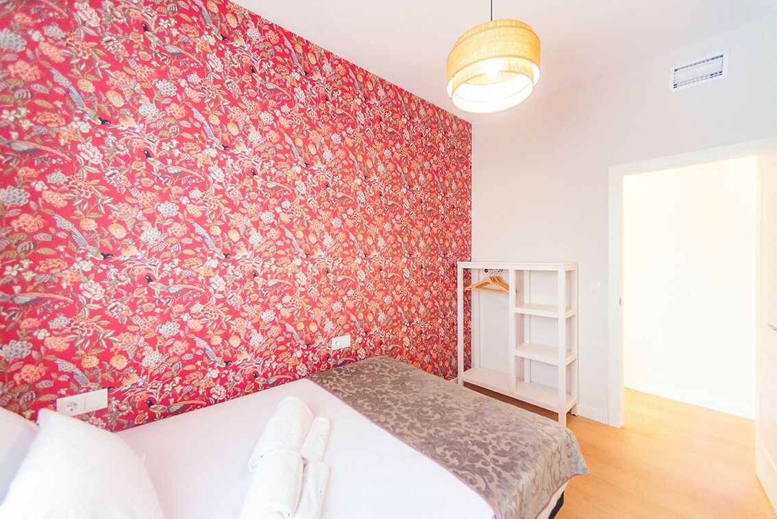 A bedroom with a floral wallpaper, a bed with a grey and white patterned bedspread, a hanging light fixture, and a white wardrobe.