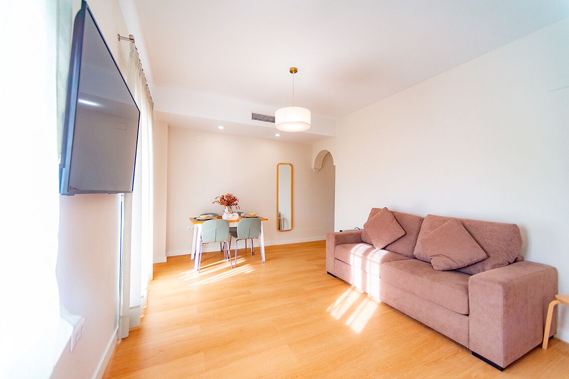 Un salon moderne avec un plancher en bois, un canapé rose, une table à manger avec des chaises, un téléviseur fixé au mur et un luminaire suspendu.