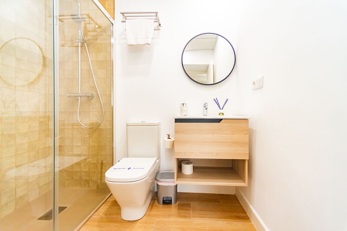 Un baño moderno con cabina de ducha de vidrio, inodoro blanco, tocador de madera con espejo y un pequeño cubo de basura.