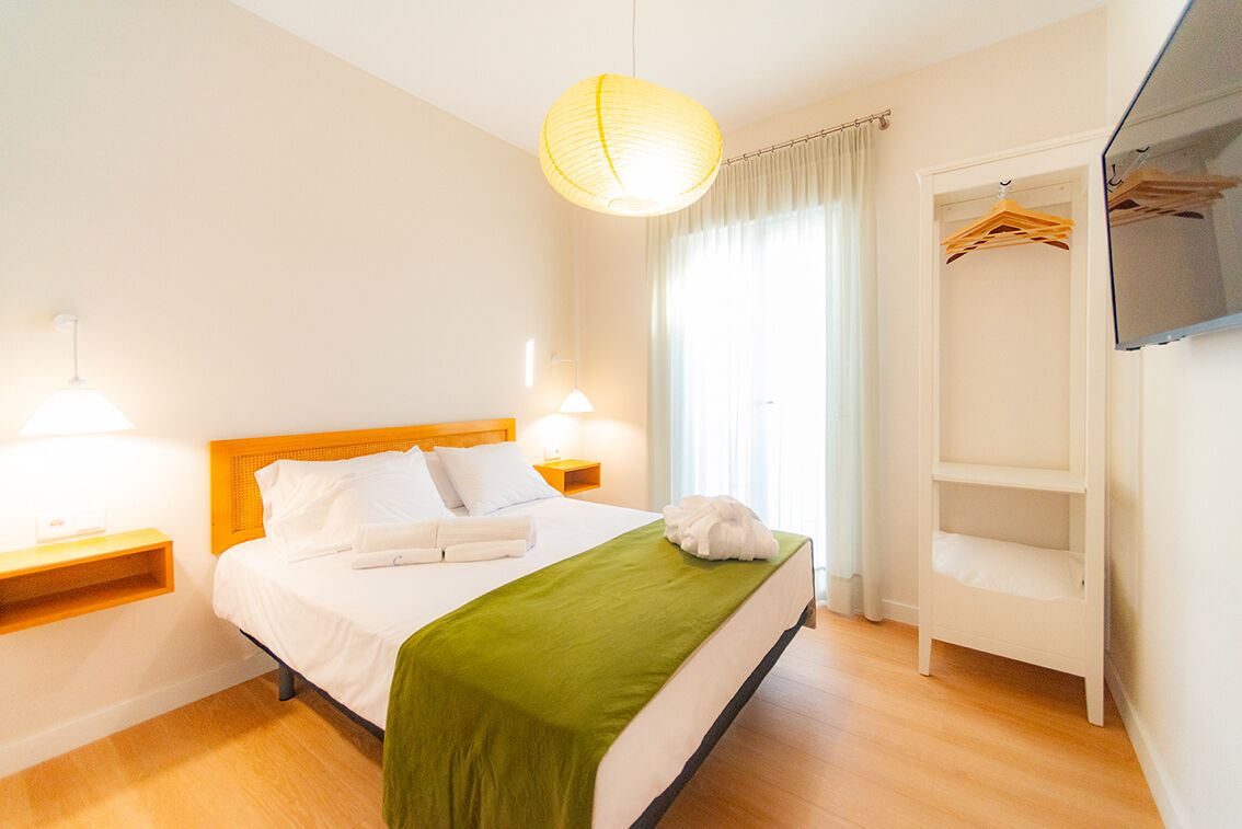 A neatly arranged hotel room with a bed, bedside lamps, a hanging light fixture, a wardrobe, and a television.