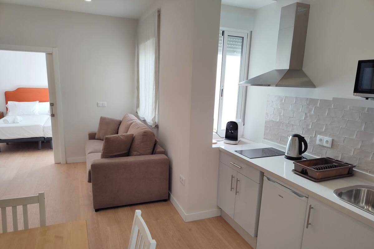A modern kitchen with a built-in oven, a kettle, and a sink. There is a dining area with a wooden table and chairs. A sofa is placed in the living area, and a bed is visible in the adjacent room.