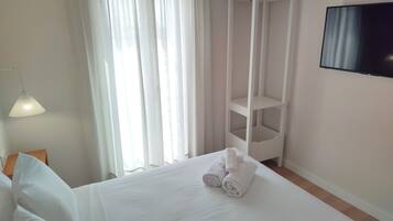 Un lit bien fait, avec du linge de lit blanc et des serviettes pliées, une table de chevet blanche, une lampe, un téléviseur fixé au mur et une fenêtre ornée de rideaux légers.