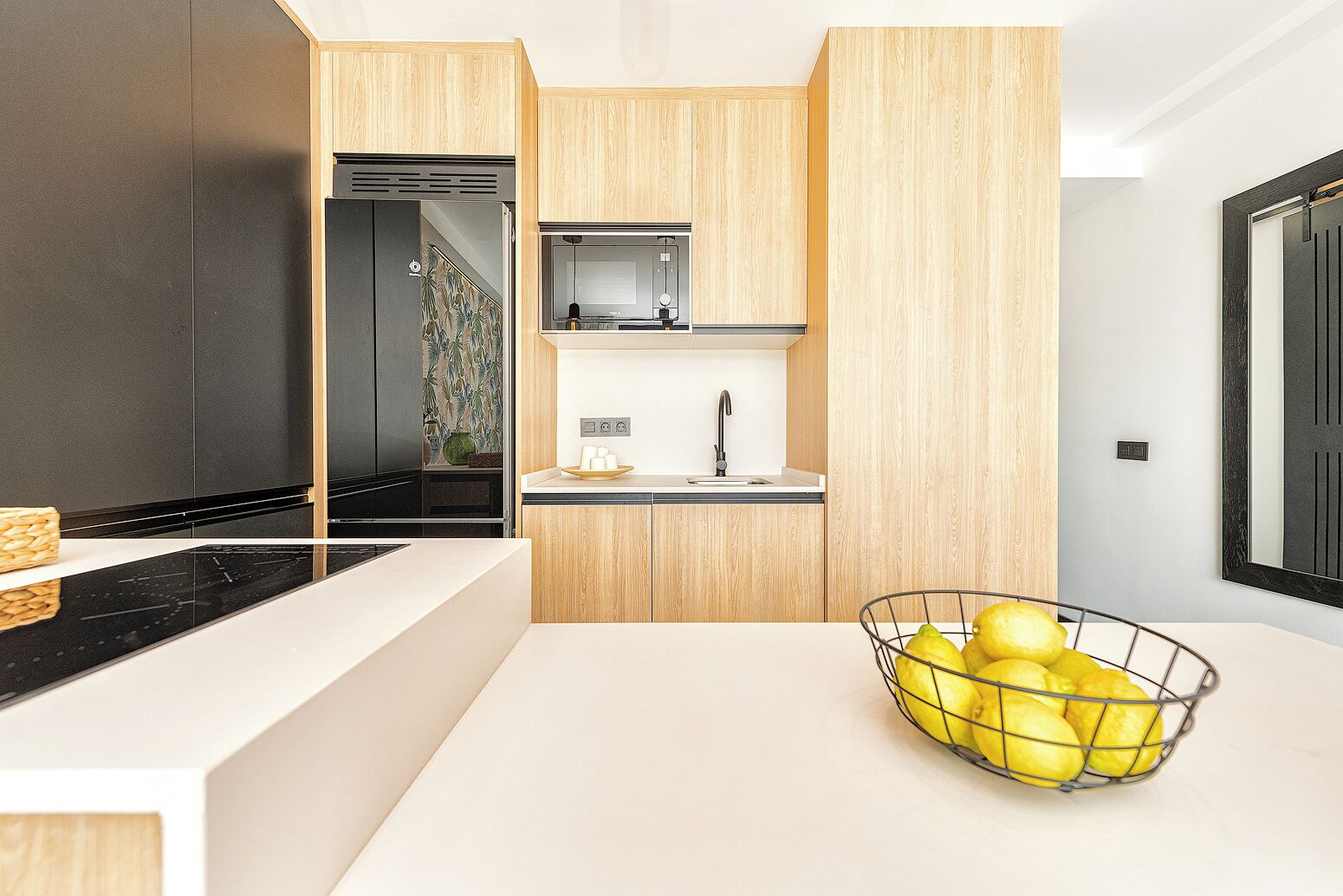 Cocina moderna con encimera blanca, salpicadero negro y armarios de madera. Hay un fregadero con grifo y una pequeña ventana encima. Una cesta de limones está sobre la encimera.