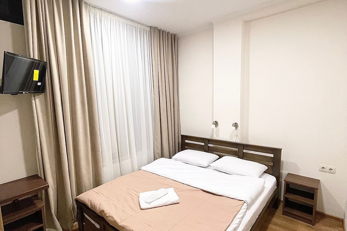 A neatly made bed with a brown bedspread and white pillows, a wooden nightstand with a lamp, and a television mounted on the wall.