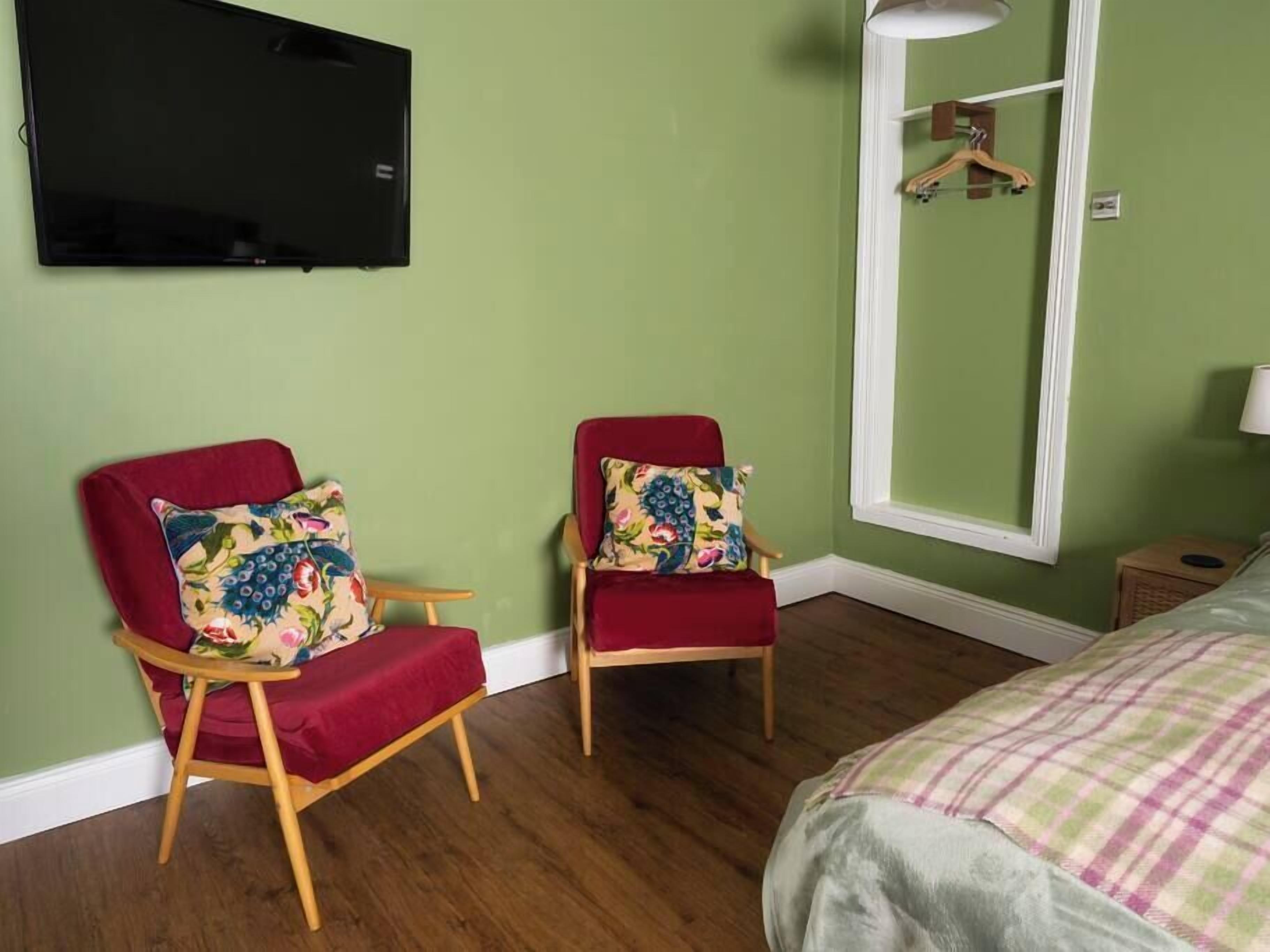 Ein Zimmer mit zwei roten Sesseln, einem Bett mit kariertem Überwurf, einem an der Wand befestigten Fernseher und einem Spiegel mit Holzrahmen.