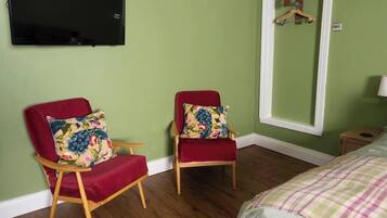 Ein Zimmer mit zwei roten Sesseln, einem Bett mit kariertem Überwurf, einem an der Wand befestigten Fernseher und einem Spiegel mit Holzrahmen.