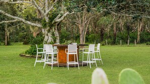 A wooden outdoor dining area with a round table and four white chairs, surrounded by lush greenery and trees.