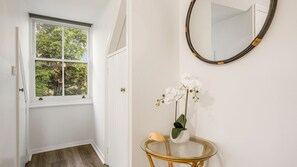 A hallway with a white door, a window with a view of trees, a round mirror on the wall, and a small table with a potted orchid.