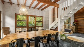 A dining area with a wooden table and chairs, large windows with a view of greenery, a hanging light fixture, and a staircase with wooden railings.