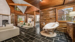 A log cabin interior with a stone-tiled floor, a white sofa, a wicker chair, and a kitchen area visible through an open doorway.