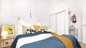 Ein ordentlich eingerichtetes Schlafzimmer mit Bett, Kissen, Nachttisch mit Lampe, gerahmtem Bild und Feuerlöscher an der Wand.