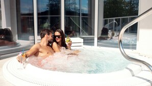 Un couple se détend dans un bain à remous, un verre à la main, entouré de grandes fenêtres offrant une vue dégagée sur l'extérieur.