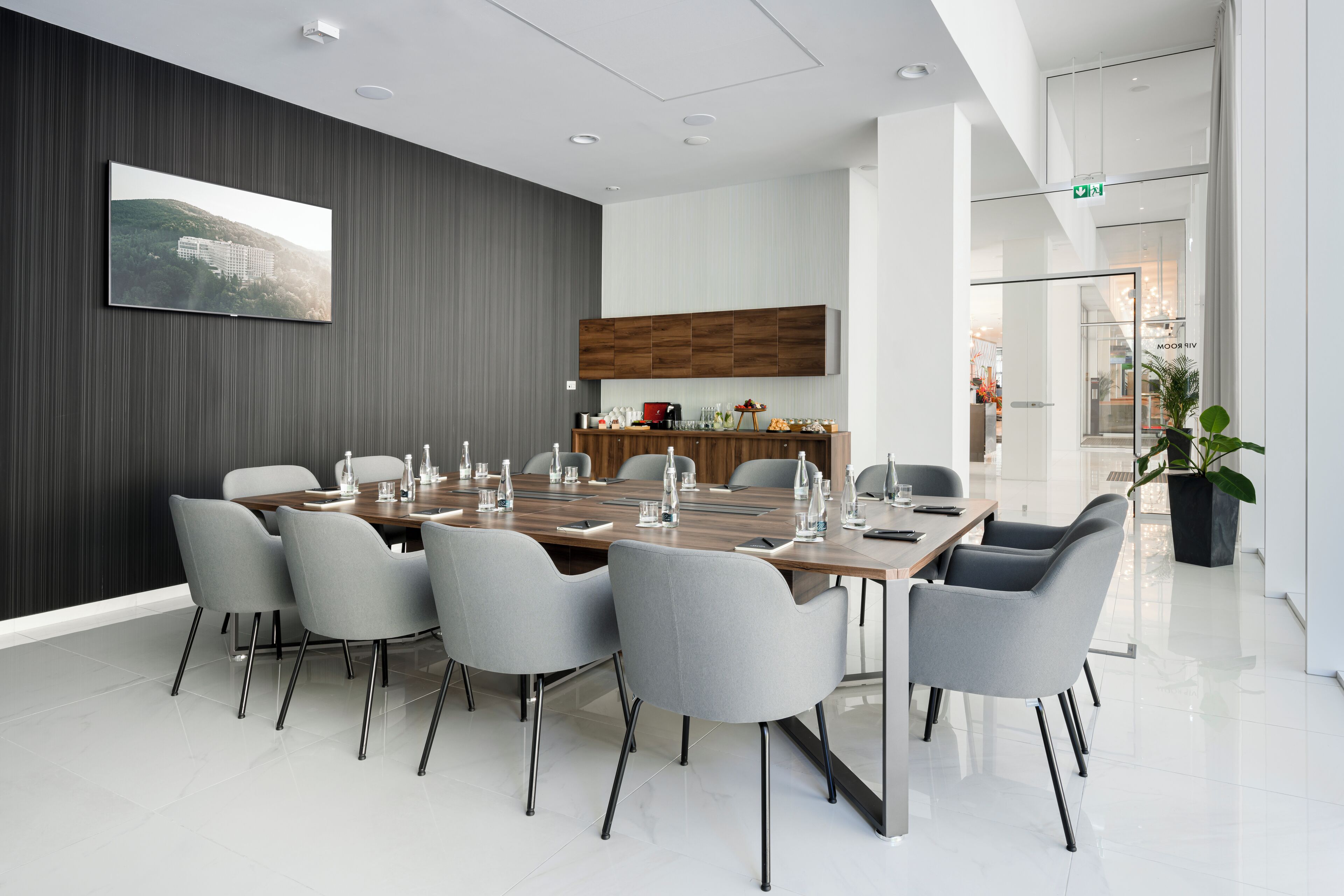 A modern conference room with a long table, grey chairs, a wooden cabinet, and a large flat-screen TV on the wall.