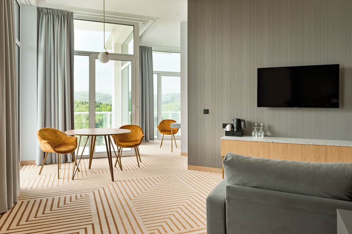 Una habitación de hotel moderna con un ventanal grande, un televisor de pantalla plana, un sofá y un área de comedor con dos sillas.