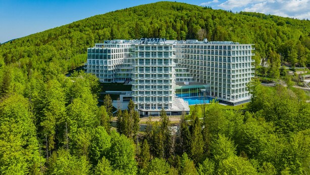 Un hôtel moderne doté d’une façade en verre, d’une piscine, et entouré d’une végétation luxuriante et d’une colline boisée.