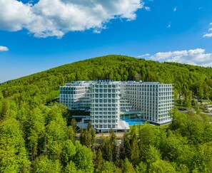 A modern hotel with a glass facade, a pool, and is surrounded by lush greenery and a forested hill. - Hotel Crystal Mountain (Wisla)