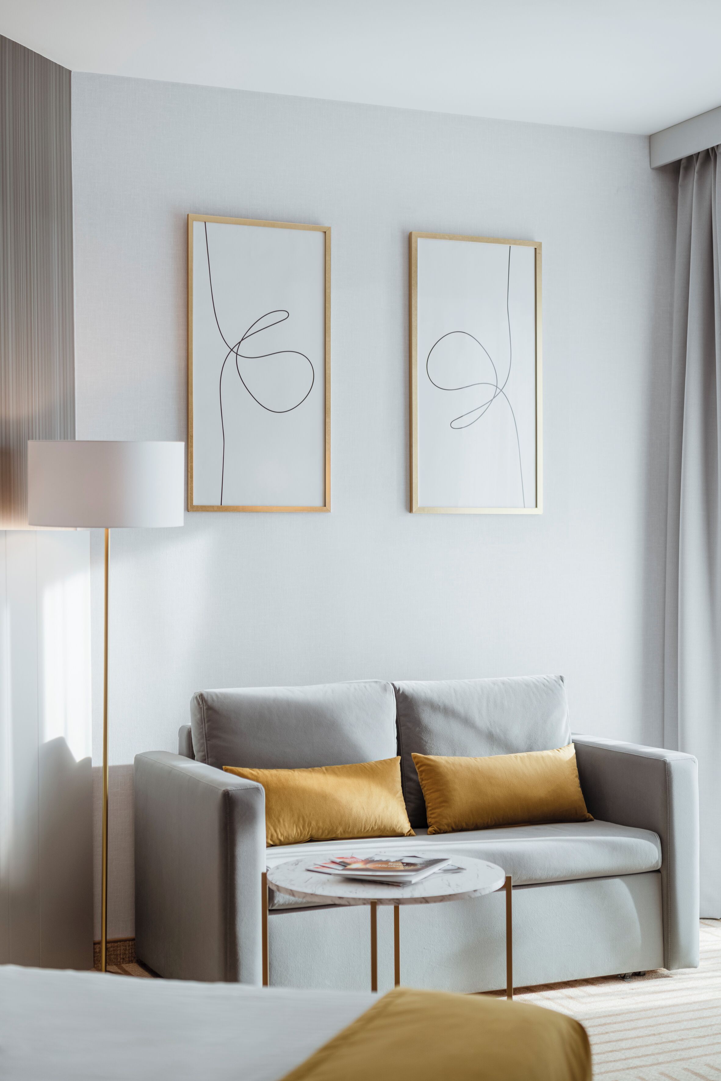 A modern hotel room with a grey sofa, a small round table, and two abstract art pieces on the wall.