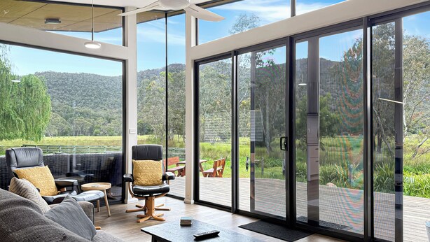 Una sala de estar moderna con un amplio ventanal corredera que da a una terraza exterior, un ventilador de techo y vistas de un paisaje exuberante.