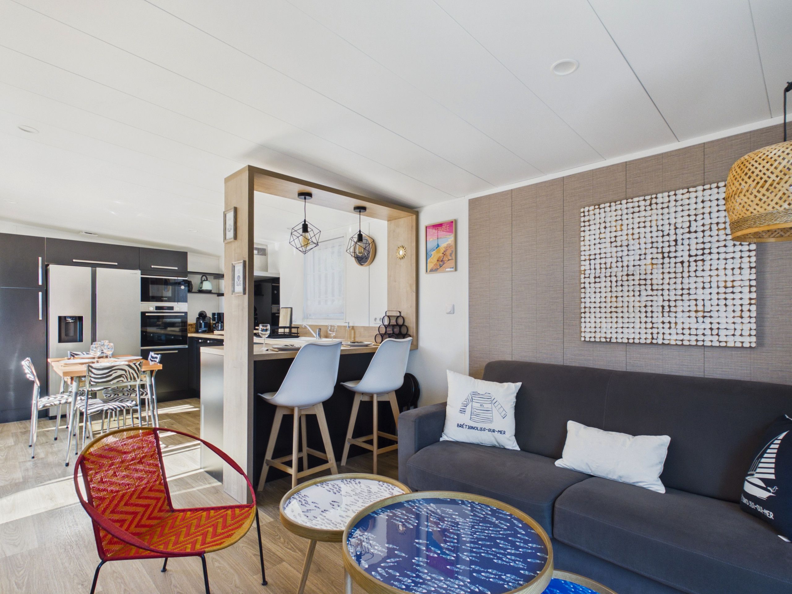 Una sala moderna con un sofá gris, un comedor con una mesa y sillas de madera, y una cocina visible a través de un arco.