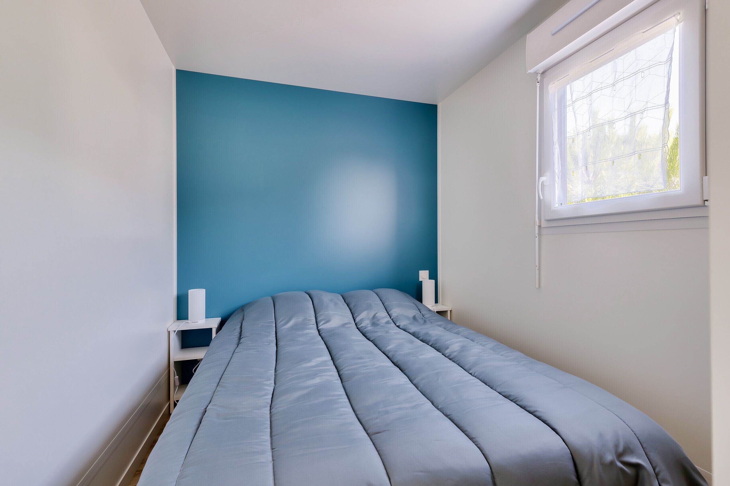 Un lit simple avec un matelas gris dans une chambre dont un mur est peint en bleu et qui possède une fenêtre avec des stores blancs.