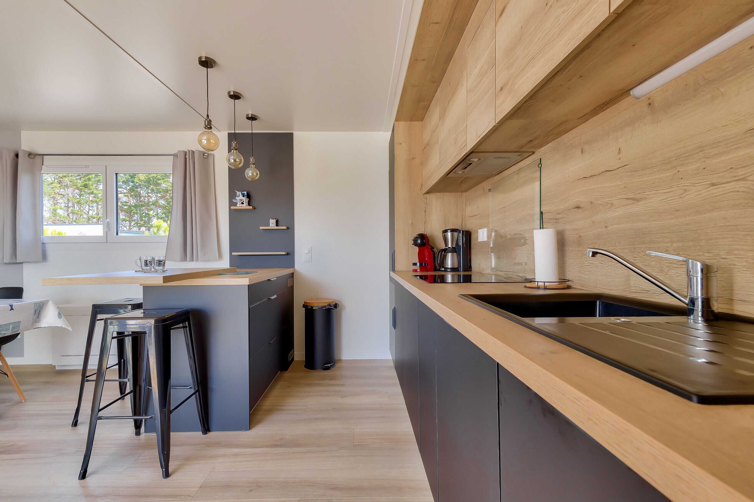 Une cuisine moderne avec un plan de travail en bois, un évier et une machine à café. On y trouve une fenêtre avec des rideaux, une poubelle et un tabouret.