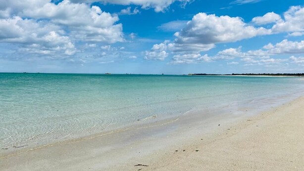 Een strand met helder turquoise water en een zandstrand, onder een blauwe lucht met losse wolken.
