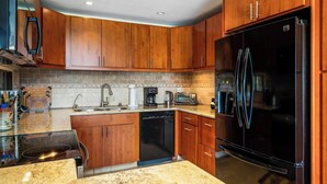 A modern kitchen with wooden cabinets, a black refrigerator, a dishwasher, and a granite countertop. - Keauhou Punahele A103 | Ground Floor Condo with Split AC and Oceanviews (Kailua-Kona)
