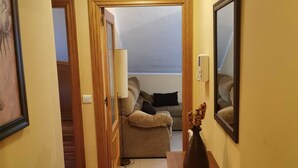 A hallway with wooden flooring, a sofa in the background, a vase on a table, and a ceiling light. - Apartamento Cinema (Cangas de Onís)
