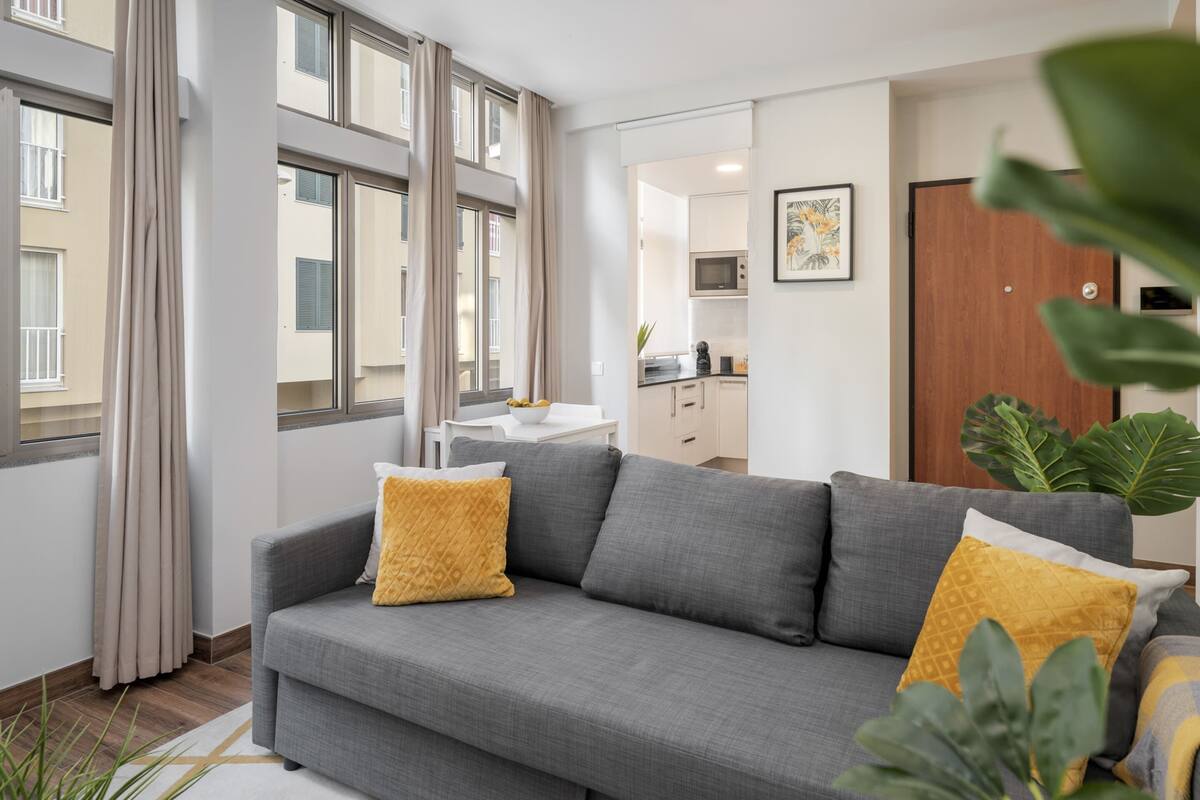 A modern living room with a grey sofa, yellow cushions, and a view of the kitchen in the background.
