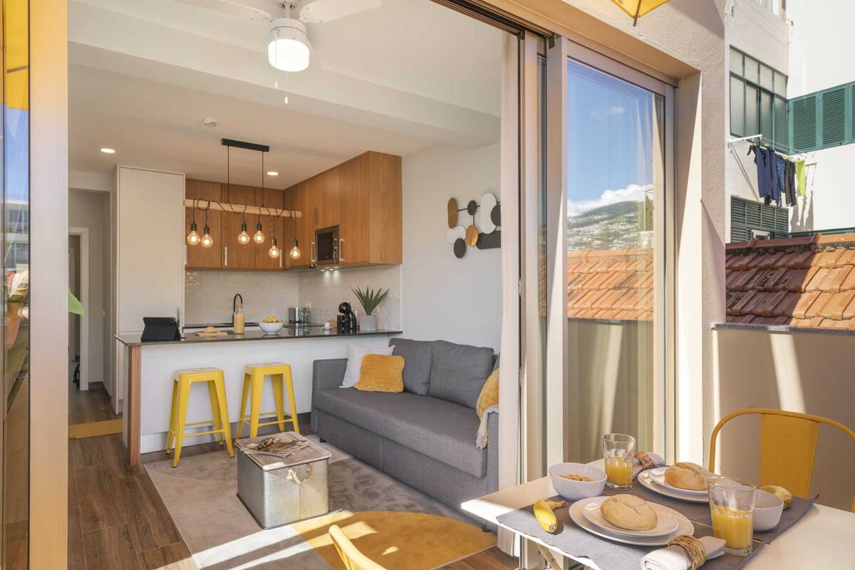 A modern kitchen with wooden cabinets, a grey sofa, and yellow stools. There's a dining area with a table set for breakfast, including plates, glasses, and a banana.