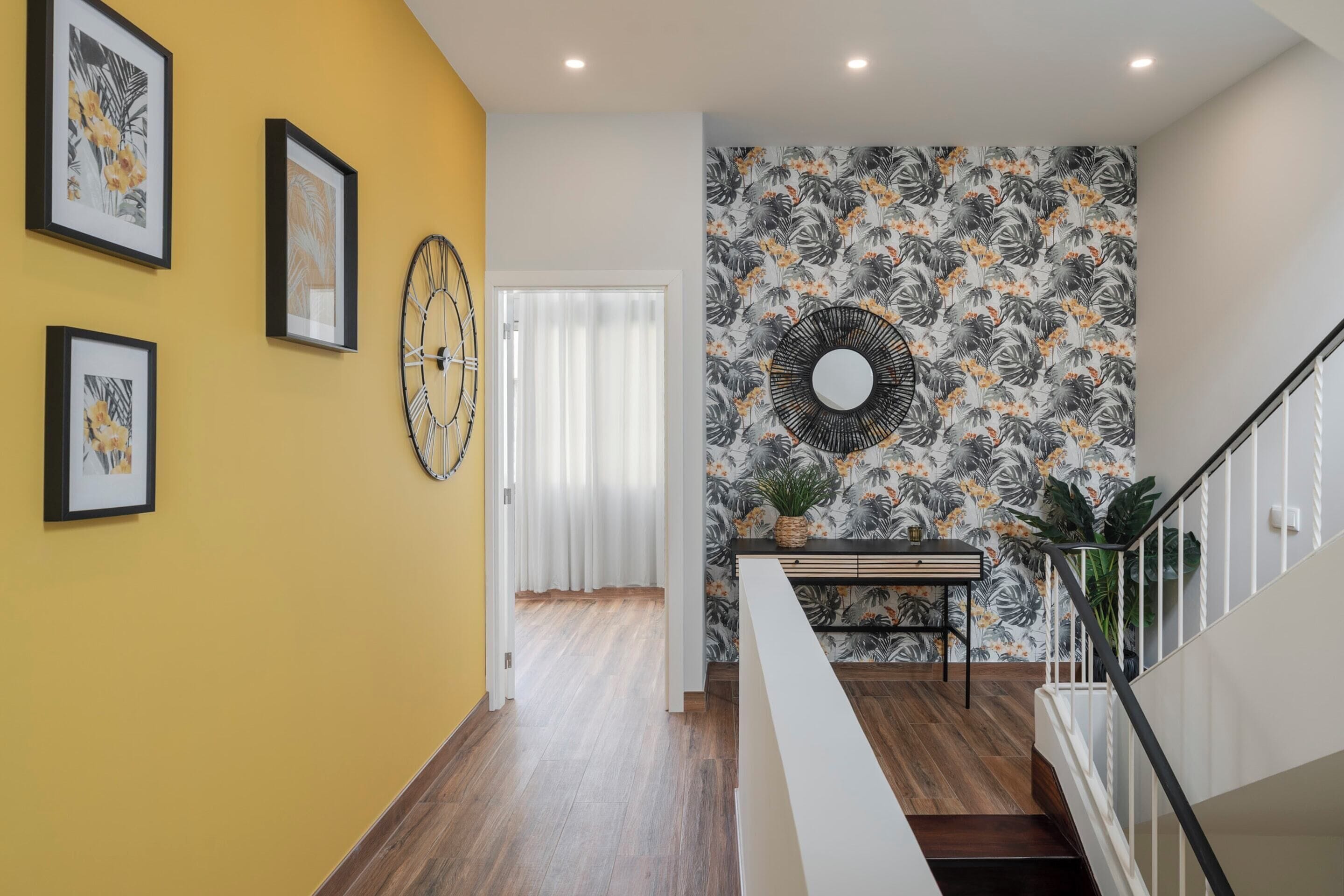 A hallway with a yellow wall, framed artwork, a round mirror, and a staircase with a railing.