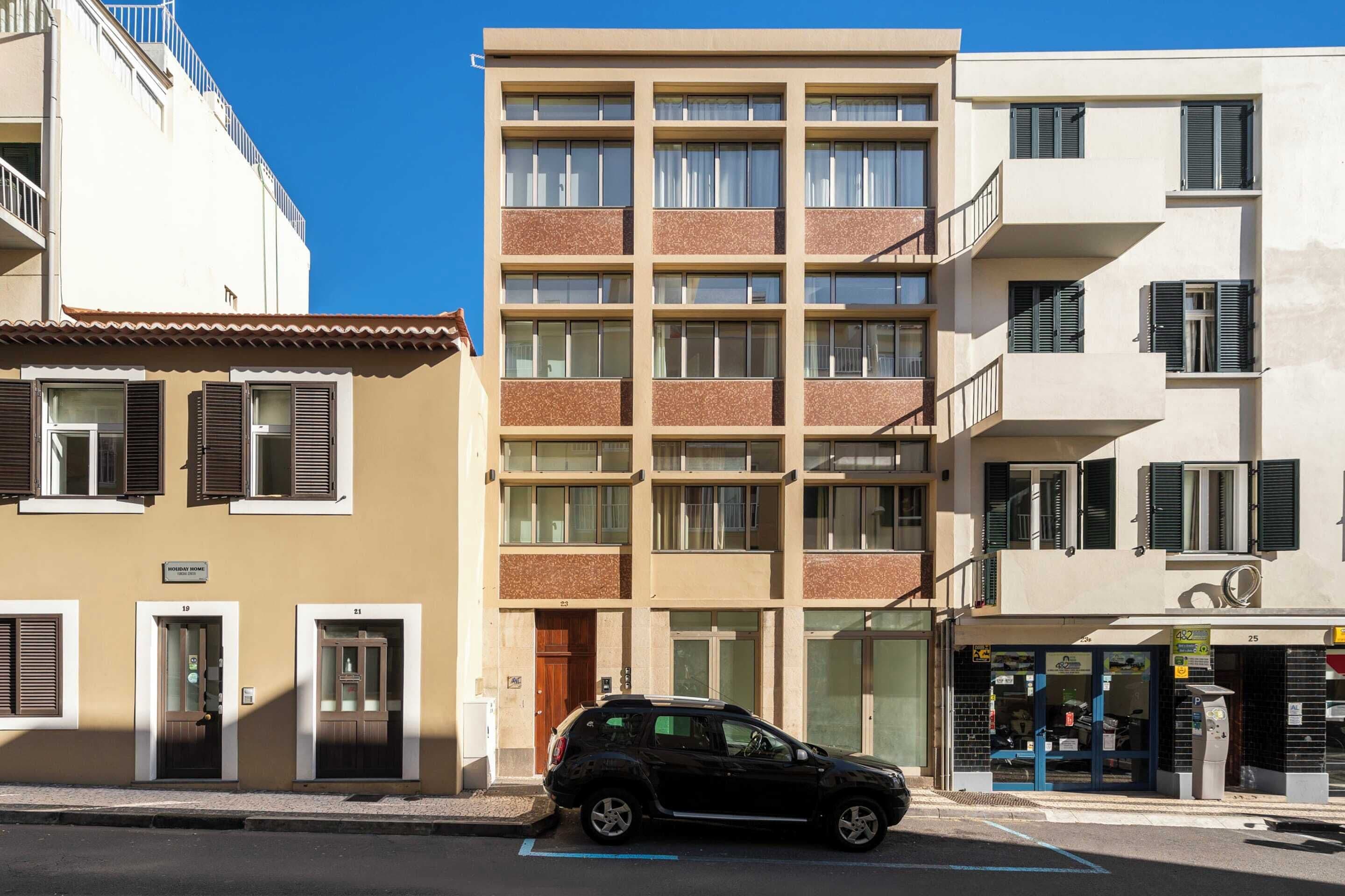 Ein mehrstöckiges Gebäude mit Eingang im Erdgeschoss, ein geparktes Auto davor und ein sichtbarer Laden mit Schaufenster.