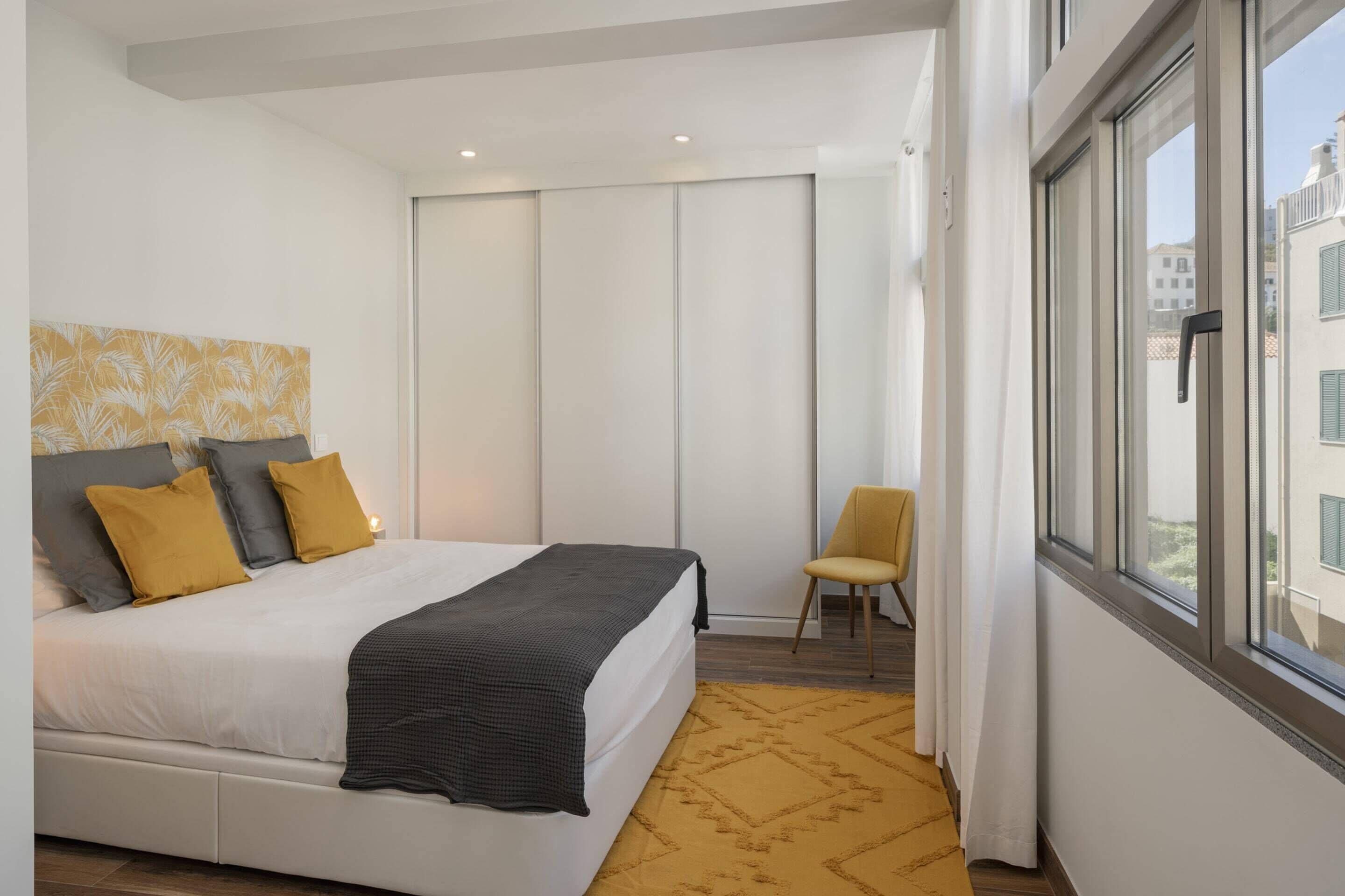 A modern bedroom with a large bed, a chair, and a view of buildings through a window.