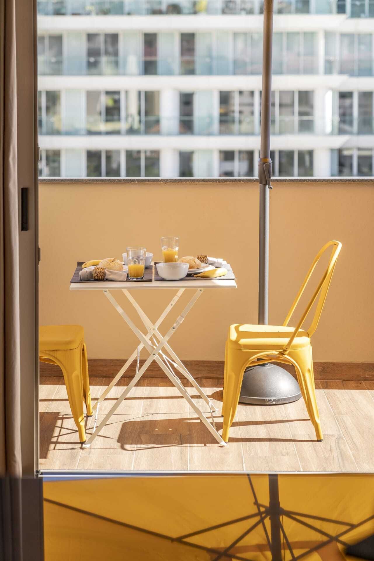 Parvekkeella on aamiaispöytä, keltaiset tuolit ja näkymä moderniin rakennukseen.