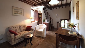 A cozy living room with a floral sofa, a white armchair, a wooden coffee table, and a dining table with a vase of flowers. - Castello di Montalto - San Martino (Castelnuovo Berardenga)