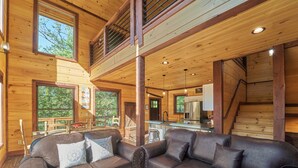 A wooden cabin interior with a dining area, kitchen, and a large window overlooking greenery.