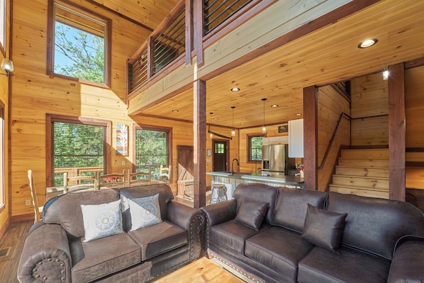 A wooden cabin interior with a dining area, kitchen, and a large window overlooking greenery.