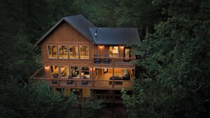 A two-story wooden cabin with multiple windows and a balcony, surrounded by dense forest.
