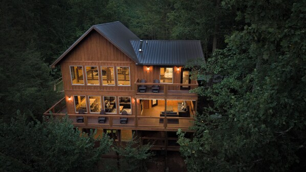A two-story wooden cabin with multiple windows and a balcony, surrounded by dense forest.