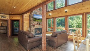 A cozy cabin interior with a stone fireplace, wooden walls, and large windows offering a view of the outdoors.