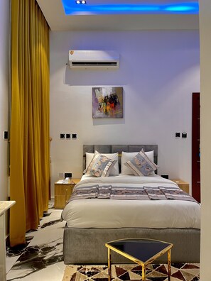 A modern bedroom with a bed, bedside tables, an air conditioning unit, a ceiling light, and a wall-mounted artwork. - Lisa Suites (Abuja)