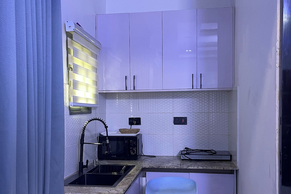 Une cuisine compacte avec des meubles blancs, un four à micro-ondes, un évier et un petit tabouret.